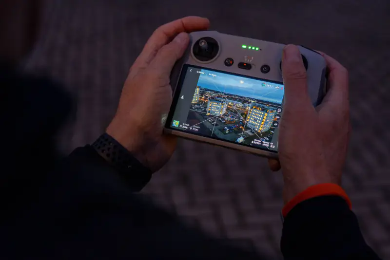 Handen om de DJI-controller in de schemering in Enschede; op het scherm het live beeld van de drone boven verlichte kantoren