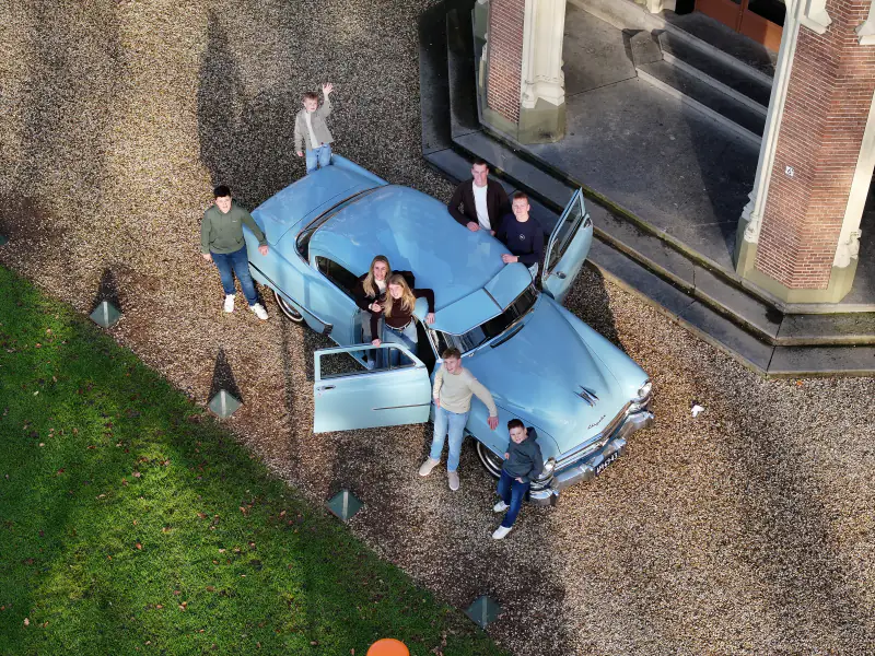 Familie rond een lichtblauwe oldtimer bij de trap van een landhuis, recht van boven met de drone; een van de kinderen zwaait