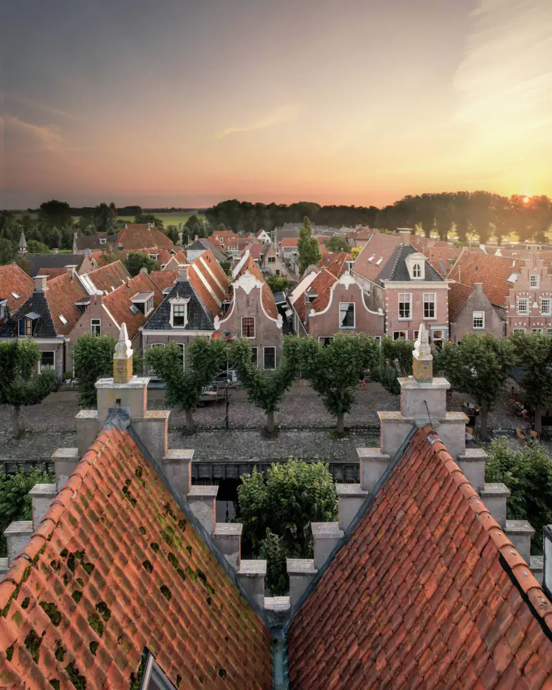 Daken, trapgevels en een bomenrij langs de gracht van een oud stadje bij zonsondergang, vanuit de drone