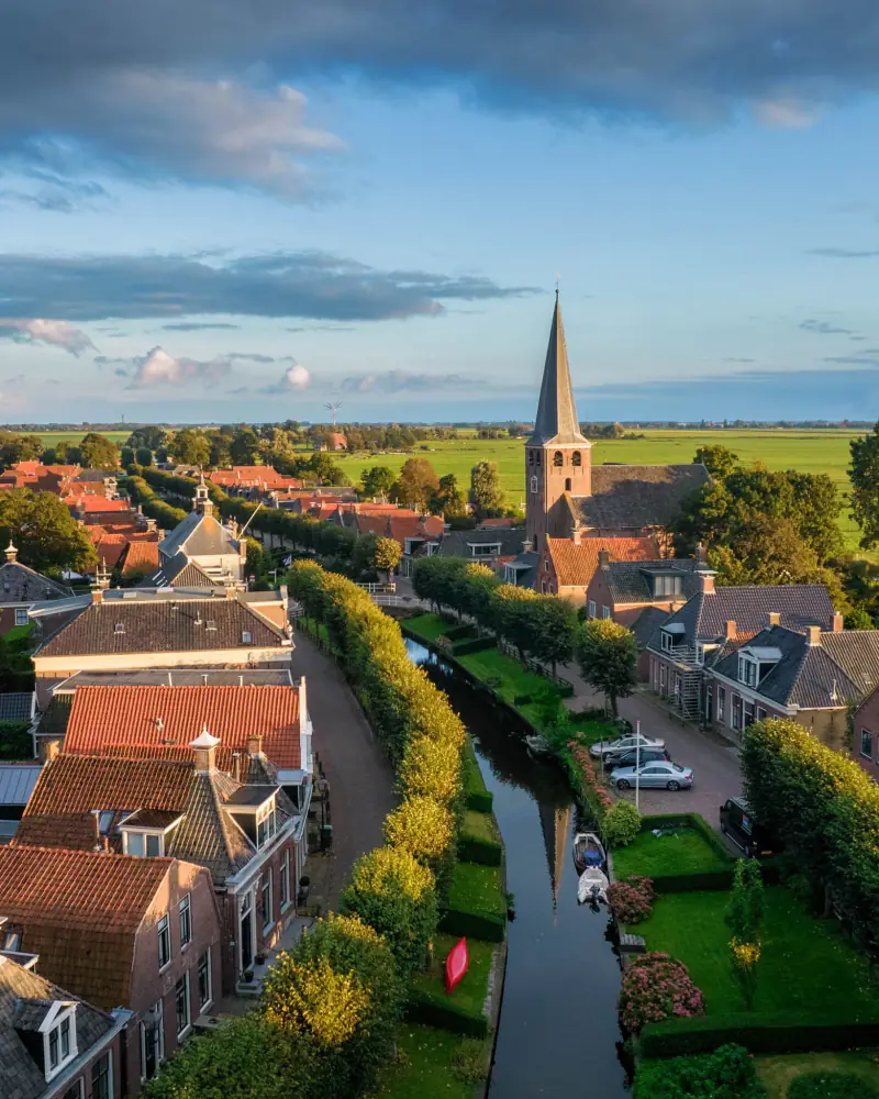 Kerkdorp met een gracht tussen geschoren bomen en weilanden tot aan de horizon, vanuit de drone