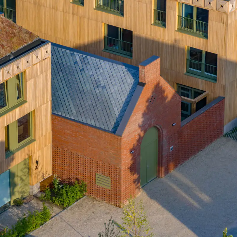 Rood bakstenen gebouwtje met leien dak en groene deur tussen houten gevels, schuin van boven in het avondlicht
