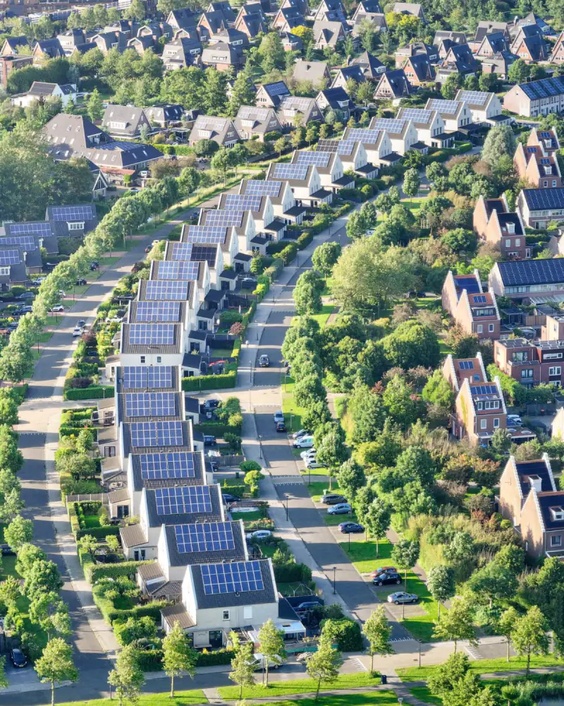 Nieuwbouwwijk van boven: rijen huizen met zonnepanelen op het dak langs lanen met bomen, in het ochtendlicht